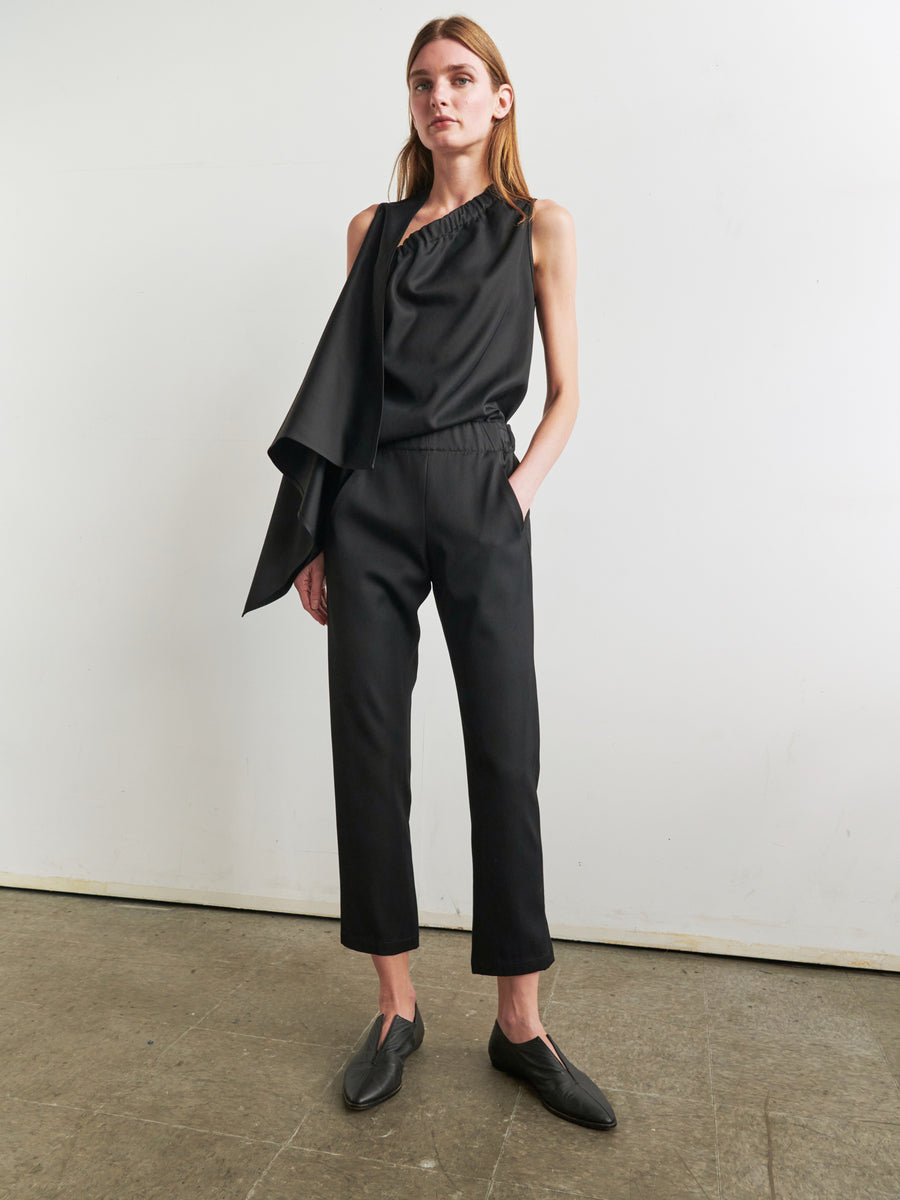 A person with long hair is standing indoors against a plain white wall. They are wearing a stylish, asymmetrical black top with a draped detail on one shoulder and the Slim Elli Pant by Zero + Maria Cornejo, which features hidden pockets and relaxed legs. One hand rests casually in their pocket, and they sport black slip-on shoes.