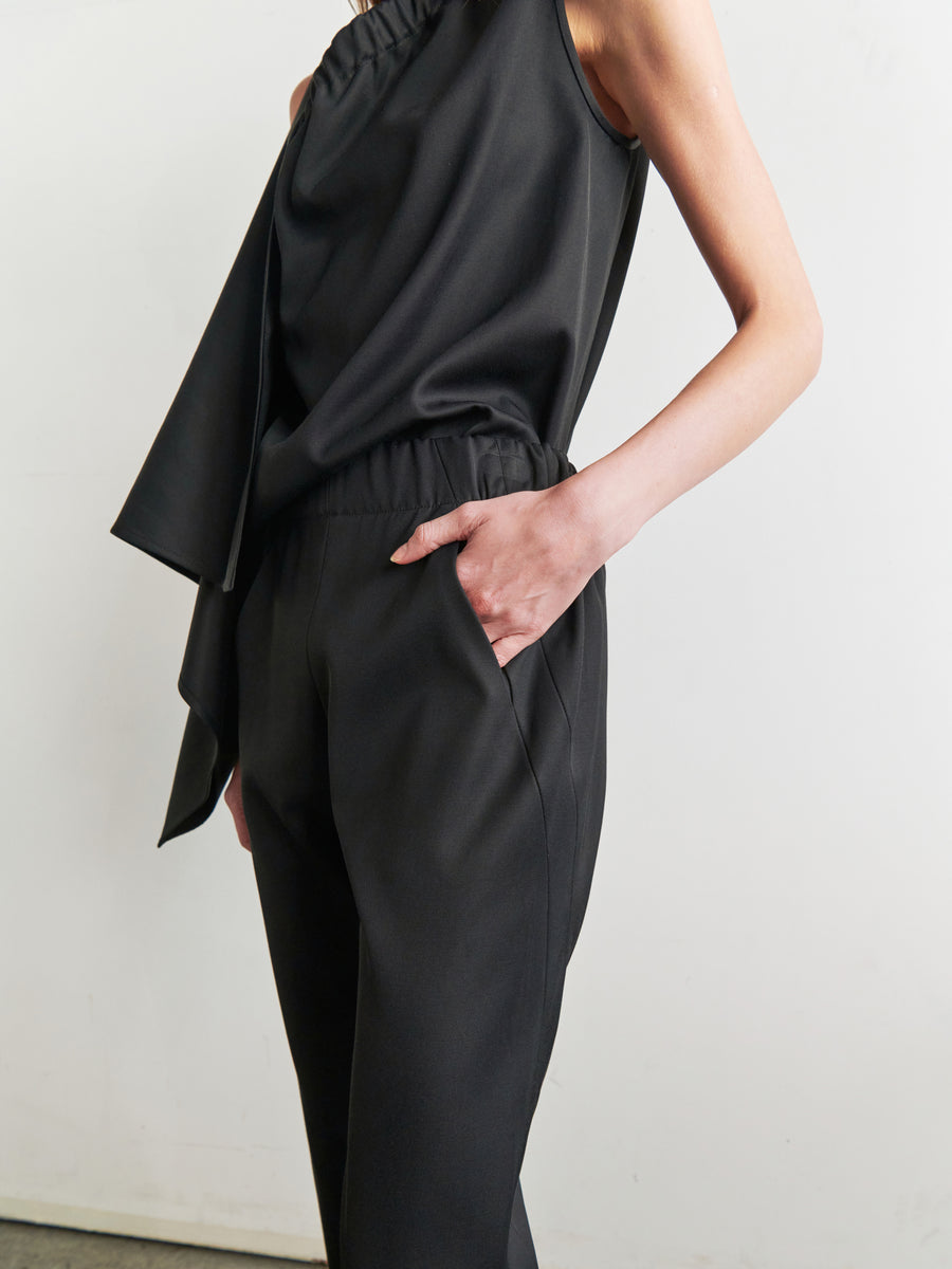 A person stands against a plain white background, wearing a sleeveless black top with an asymmetrical drape and high-waisted Slim Elli Pants from Zero + Maria Cornejo. Their left hand rests in the pocket of the relaxed-leg trousers. The overall style is minimalist and elegant.