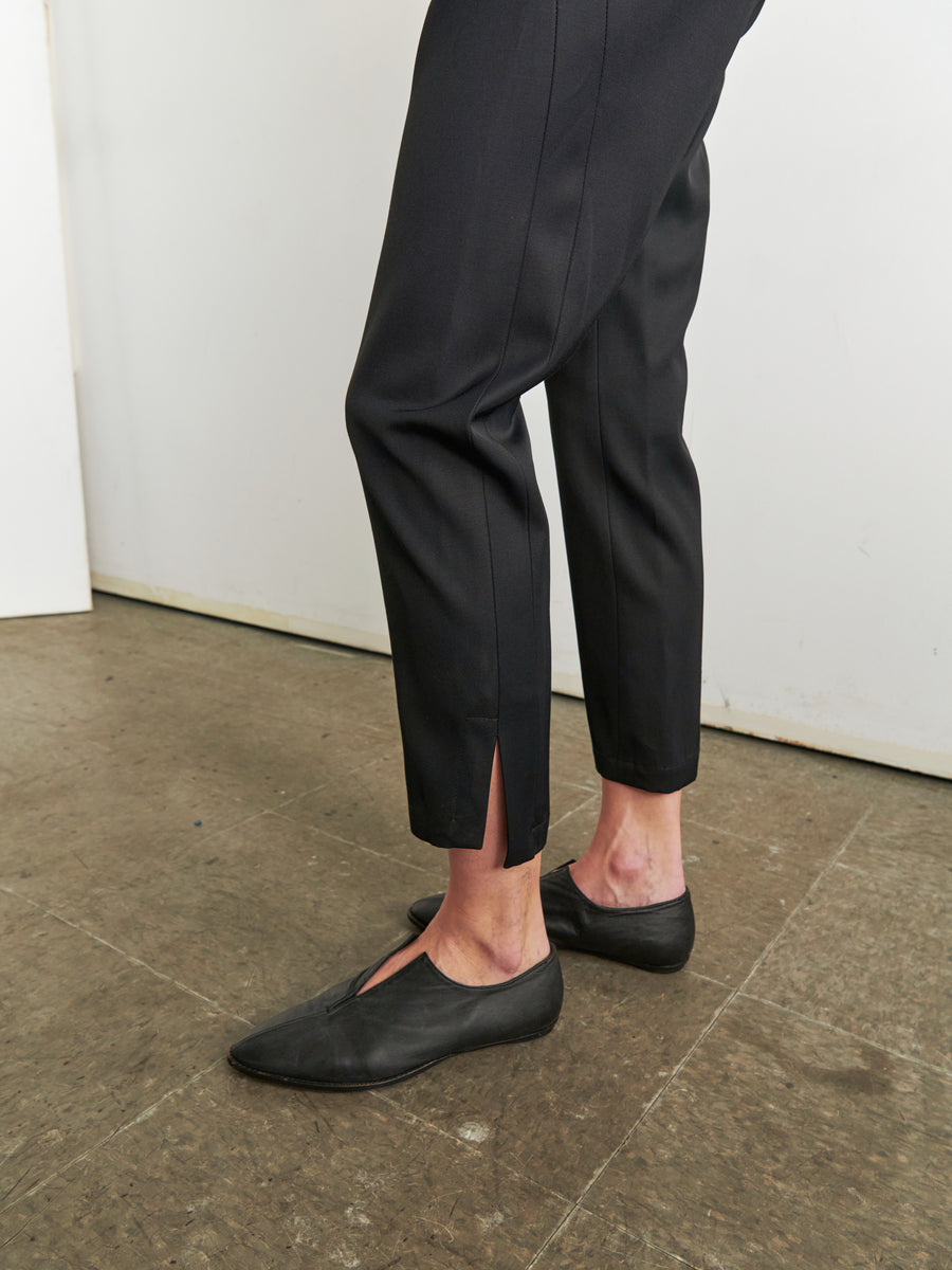 A person wearing the black Slim Elli Pant by Zero + Maria Cornejo, featuring relaxed legs and small slits at the hems, along with black slip-on shoes stands on a mottled gray floor. The lower legs and feet are visible against a white and beige background.