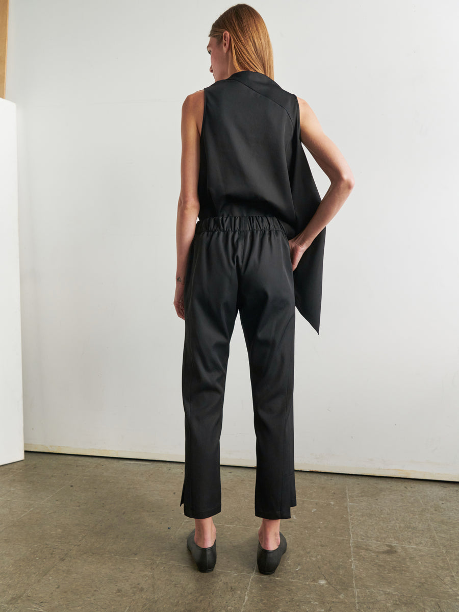 A person with long hair stands with their back to the camera, wearing a sleeveless black top featuring an asymmetrical design, Zero + Maria Cornejo's Slim Elli Pant made from relaxed leg viscose-cotton twill, and black shoes. The background is plain with a concrete floor and white wall.