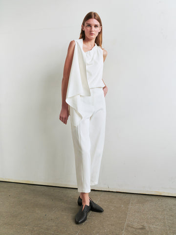 A woman with long light brown hair stands against a white wall, wearing Zero + Maria Cornejo's sleeveless asymmetric draped top and the Margot Pant in viscose-cotton twill. She pairs it with black shoes on a concrete floor.