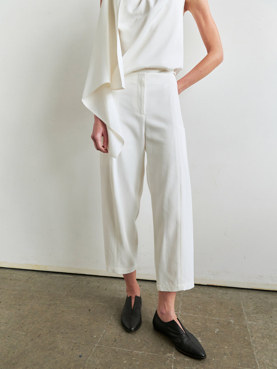 A person stands against a plain background, wearing a white asymmetrical top and the Margot Pant in white from Zero + Maria Cornejo. They have black pointed shoes and one hand resting casually by their side. The overall attire exudes a minimalist and modern aesthetic, thanks to the chic cut of the viscose-cotton twill Margot Pant.