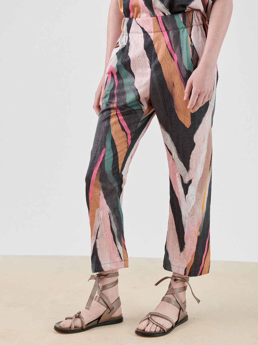 A person is wearing colorful, abstract-patterned Eko Pant and a matching top by Zero + Maria Cornejo. They are also wearing strappy, brown sandals. The background is plain and light. The scene is well-lit, showcasing the vibrant design of the clothing made from recycled polyester fabric.