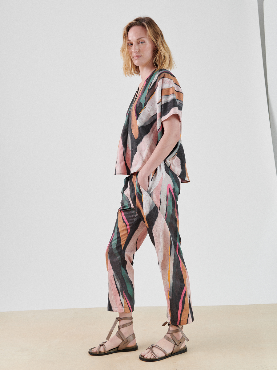 A person stands against a plain backdrop, wearing a matching set of clothing featuring an abstract metallic jacquard pattern with hues of pink, green, black, and orange. They have shoulder-length hair and are wearing flat, strappy sandals. Dressed in Zero + Maria Cornejo's Eko Pant, they look confidently at the camera.