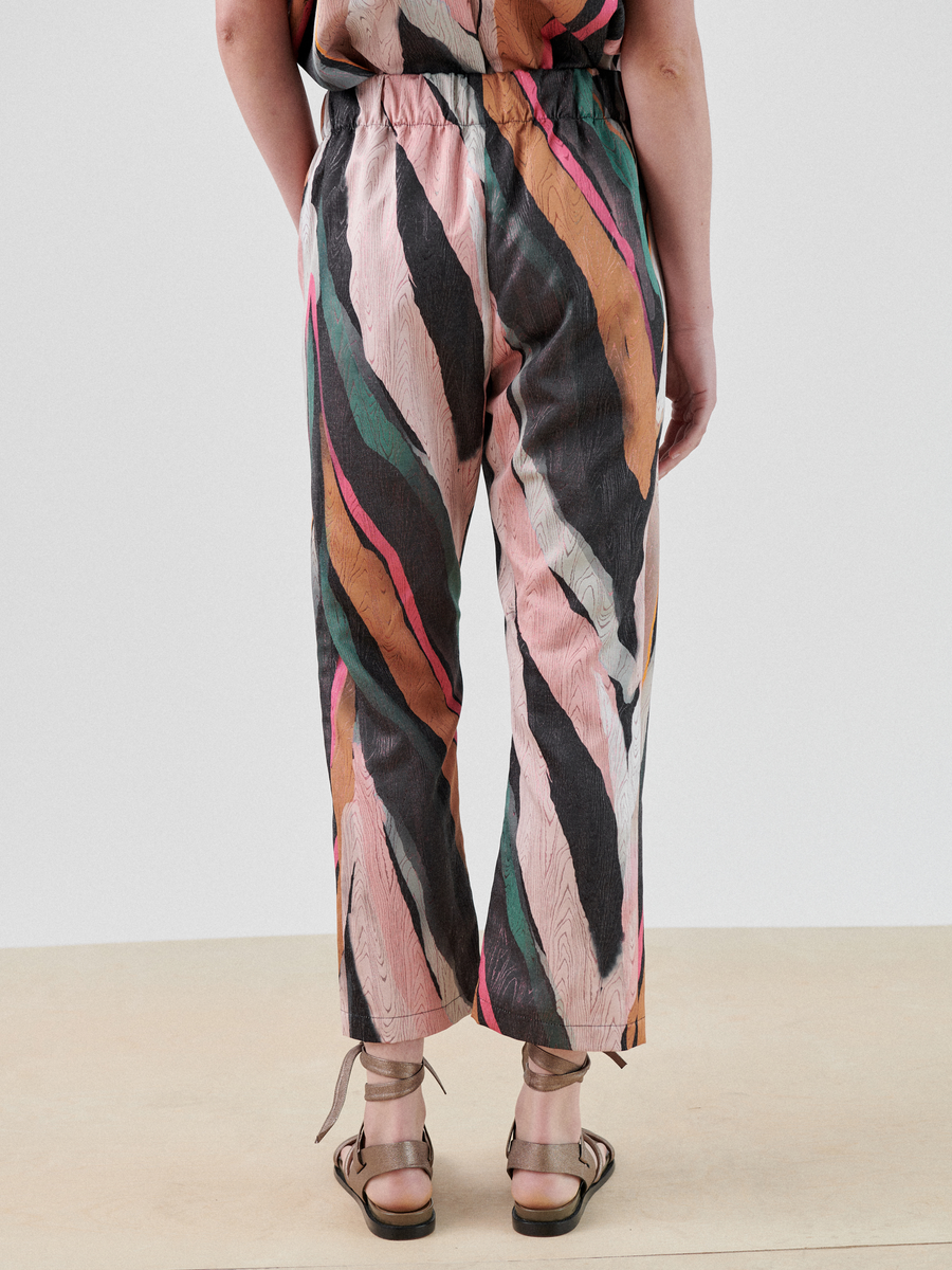 Rear view of a person standing against a plain background, wearing colorful, loose-fitting Eko Pant from Zero + Maria Cornejo made from recycled polyester with a wavy, abstract stripe pattern in shades of pink, black, green, and brown. The person is also wearing brown sandals.