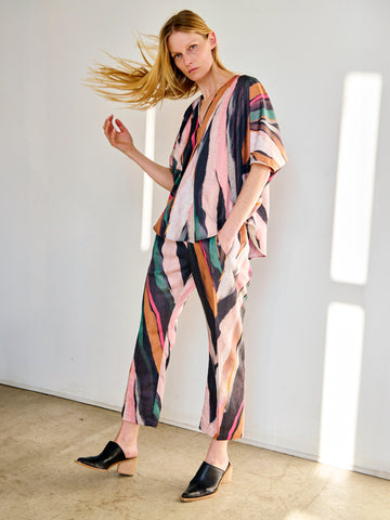 A person with long blonde hair stands in a well-lit room, flipping their hair and wearing a colorful, striped outfit consisting of a loose-fit top and matching draped Eko Pant made from recycled polyester by Zero + Maria Cornejo. They are also wearing black slip-on mules. The background is plain white.