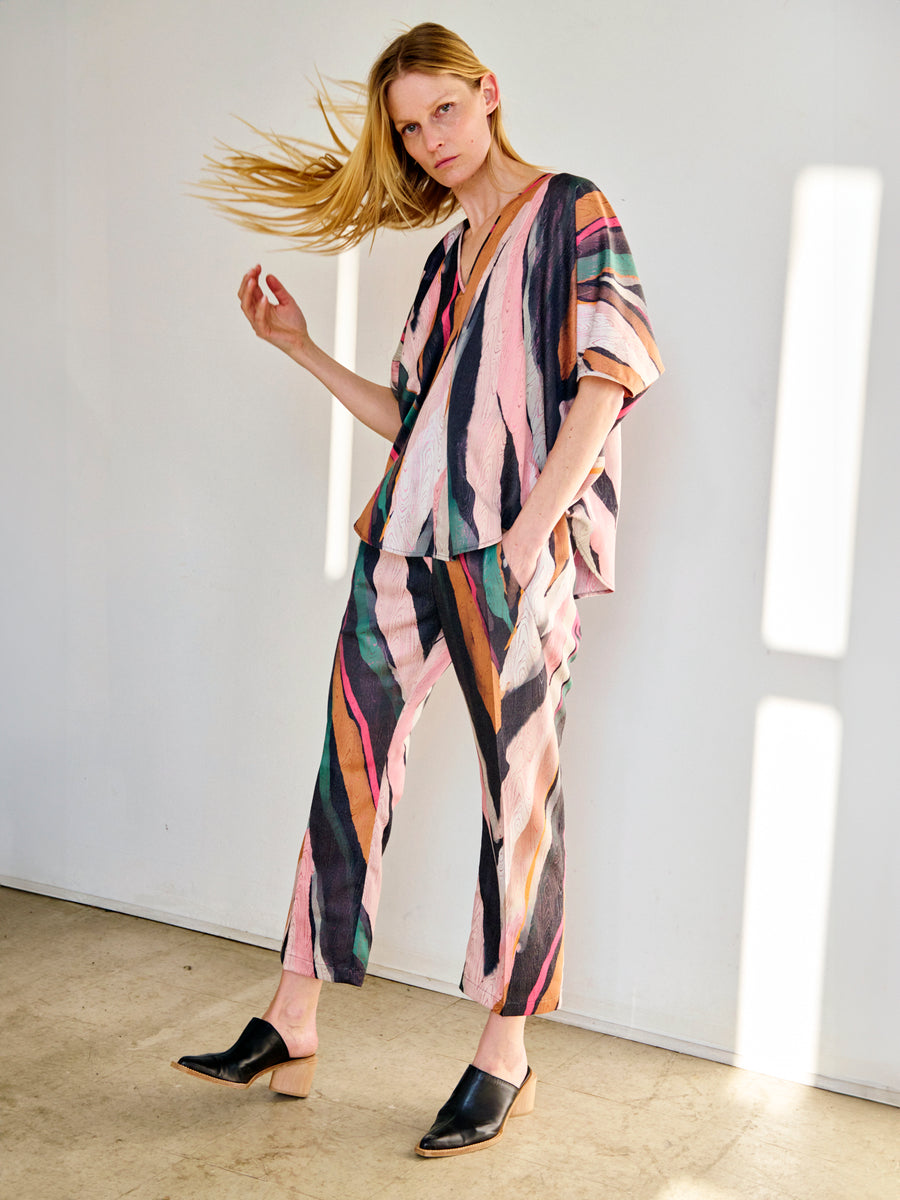 A person with long blonde hair stands in a well-lit room, flipping their hair and wearing a colorful, striped outfit consisting of a loose-fit top and matching draped Eko Pant made from recycled polyester by Zero + Maria Cornejo. They are also wearing black slip-on mules. The background is plain white.