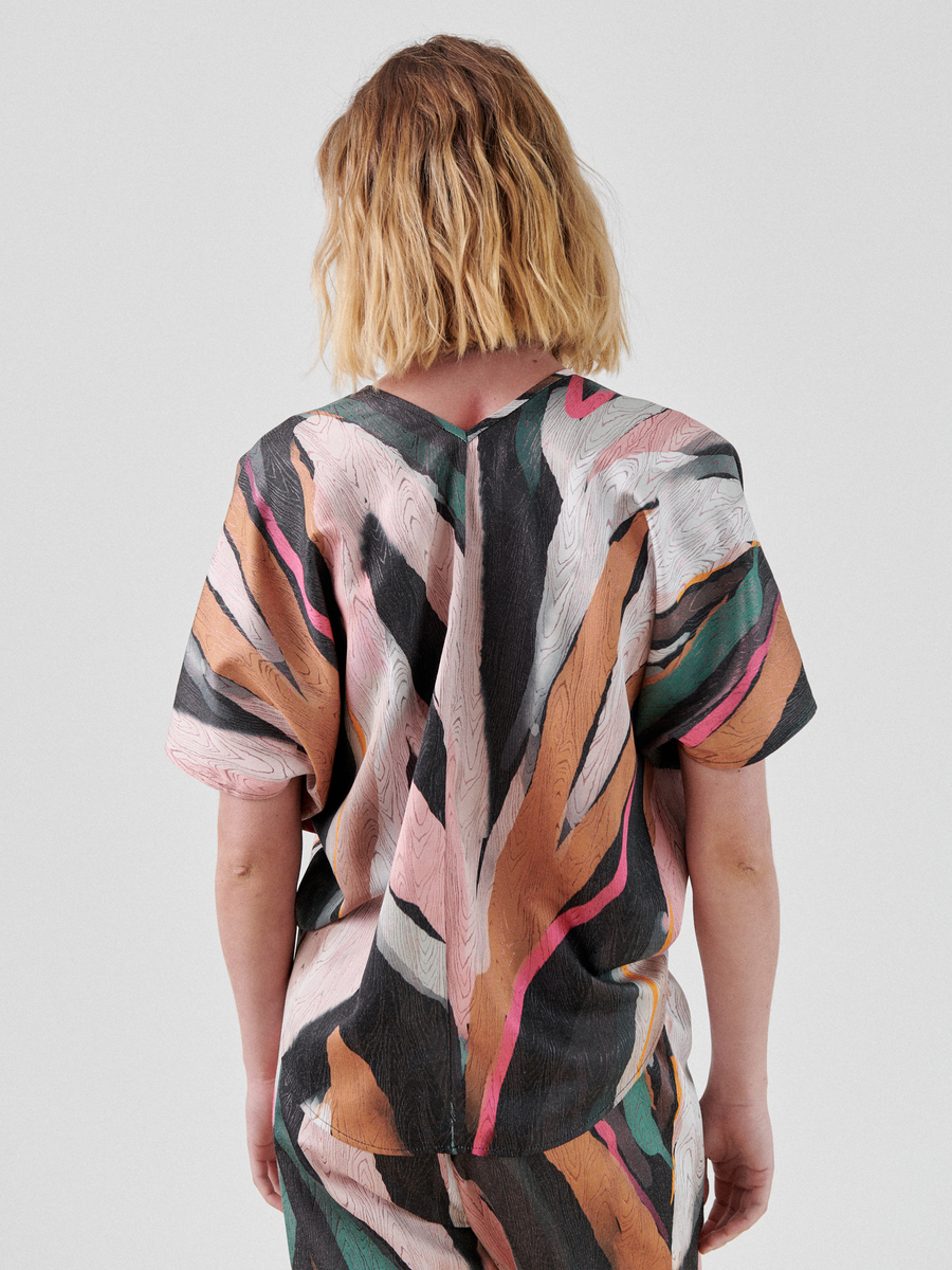 A person with shoulder-length wavy blond hair is seen from behind, wearing a colorful, abstract painterly moiré-patterned outfit consisting of the Circle Top from Zero + Maria Cornejo and matching pants. The background is a plain light gray.