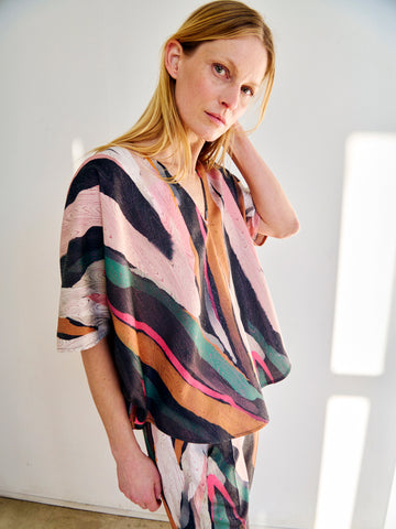 A person with long blonde hair stands against a white background, wearing the Circle Top by Zero + Maria Cornejo, a loose-fitting metallic jacquard top featuring a colorful abstract-patternly moiré pattern. The person is looking directly at the camera with a neutral expression, one hand touching their hair.