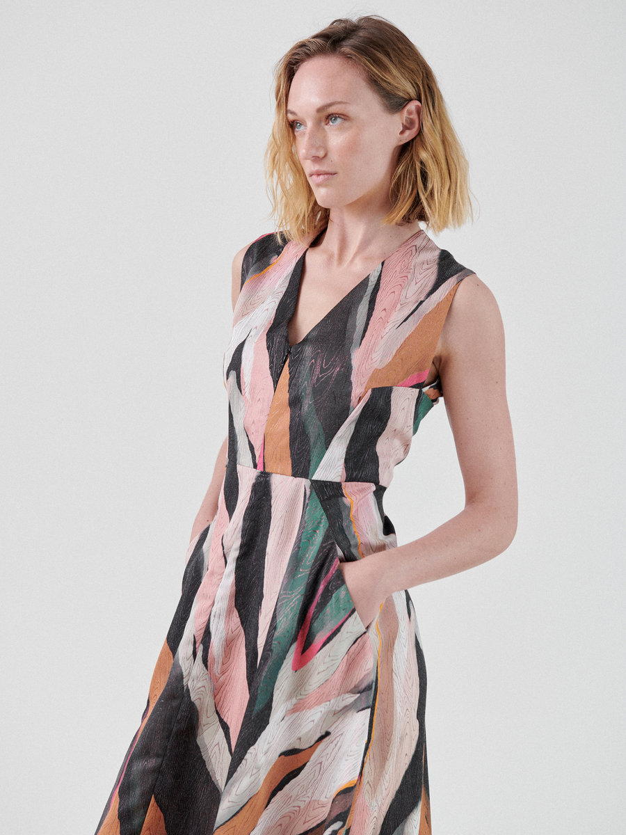 A woman with shoulder-length blonde hair wears a Sleeveless Wave Dress by Zero + Maria Cornejo, made of recycled polyester fabric, featuring a colorful abstract pattern. The dress has pockets, and she poses with one hand in a pocket, looking slightly to her left against a plain light gray background.