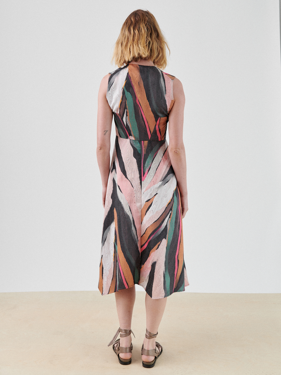 A person with shoulder-length hair stands facing away, wearing a Sleeveless Wave Dress by Zero + Maria Cornejo in bold shades of black, white, pink, orange, and green. They are also wearing strappy sandals. The background is a plain, light-colored wall and floor.