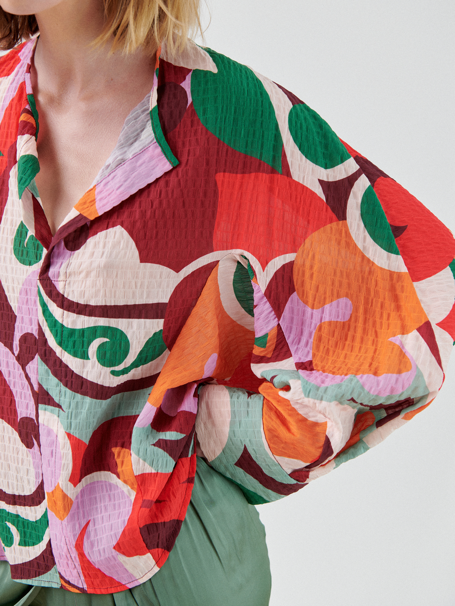 A person is shown from the shoulders to the waist wearing a Zero + Maria Cornejo Long-Sleeved Akeo Shirt with a colorful, abstract pattern in shades of red, green, orange, and purple. The shirt, made in New York, features a textured, crinkled fabric and oversized sleeves. They are also wearing green pants.