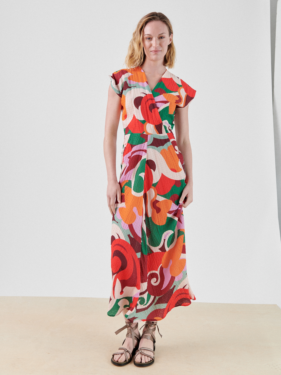 A person wearing a Zero + Maria Cornejo Wrap Omi Dress with floral and abstract designs in red, pink, green, and orange hues stands on a plain background. The vibrant wrap v-neck style makes it perfect for summer occasions. They have shoulder-length hair and are wearing brown sandals with straps.