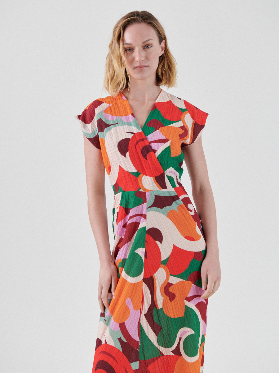 A person with shoulder-length blonde hair is wearing a colorful Wrap Omi Dress from Zero + Maria Cornejo featuring a bold, multicolor print of red, green, orange, pink, and white. Perfect for summer occasions, they stand against a plain white background, facing the camera with a neutral expression.