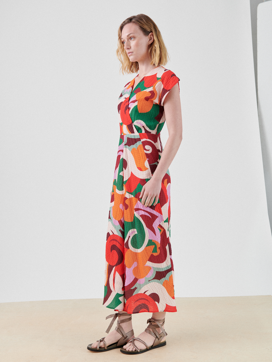 A person stands in a light-colored room wearing the Zero + Maria Cornejo Wrap Omi Dress with vibrant swirls of red, orange, green, and white. Perfect for summer occasions, the multicolor print dress features a wrap v-neck. They have shoulder-length, wavy blonde hair and are wearing light-colored sandals. The background is plain and uncluttered.