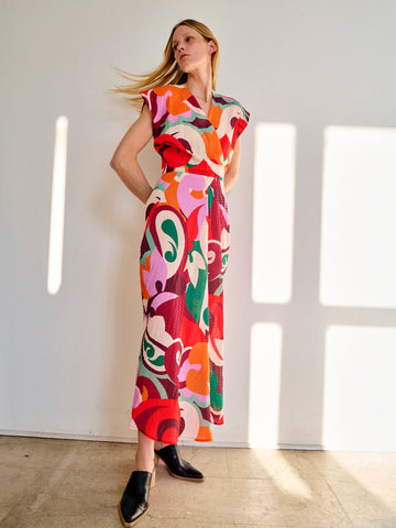 A woman stands against a white wall in a room with sunlight streaming through window panes. She is wearing the colorful, sleeveless Wrap Omi Dress with a bold, multicolor print by Zero + Maria Cornejo and black shoes. Her blonde hair is flowing, and she has a relaxed and confident posture.