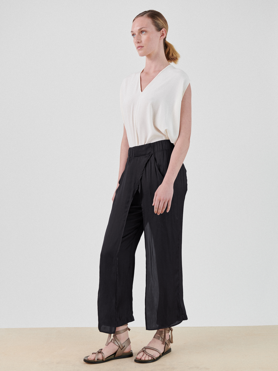 A person with pulled-back hair stands against a plain background, wearing a sleeveless white V-neck top made from recycled fibers paired with black Ido Pants by Zero + Maria Cornejo. They complete the outfit with strappy sandals. They are gazing off to the side with a relaxed expression.