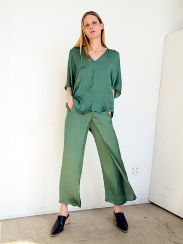 A woman with long, straight hair stands against a plain white wall, wearing a loose-fitting green blouse and matching wide-legged pants from Zero + Maria Cornejo's Ido Pant made from GRS certified polyester. She has her hands in her pockets and is wearing black slip-on shoes. The lighting is soft and natural.