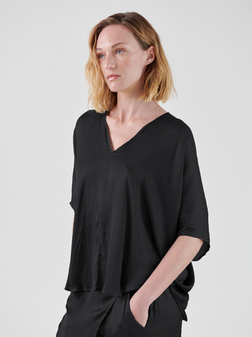 A person with shoulder-length blonde hair is standing against a plain background. They are wearing a loose-fitting, black Circle Top made from recycled polyester fabric by Zero + Maria Cornejo and have one hand in their pocket. They are gazing slightly to the side with a calm expression.