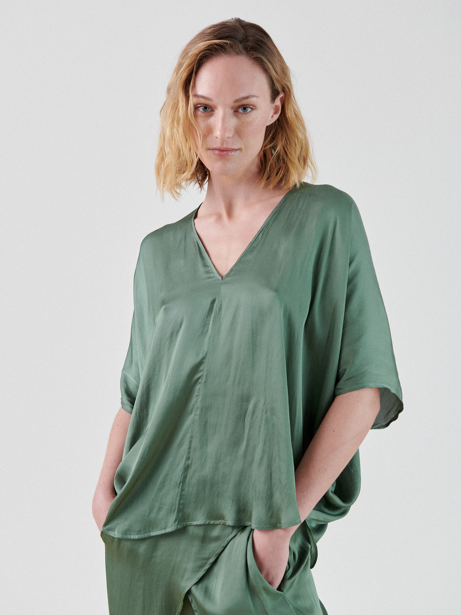 A person with shoulder-length, wavy blonde hair is wearing a loose, green V-neck Circle Top by Zero + Maria Cornejo with short sleeves. Their hands are in the pockets of matching green pants made from recycled polyester. The background is plain white.