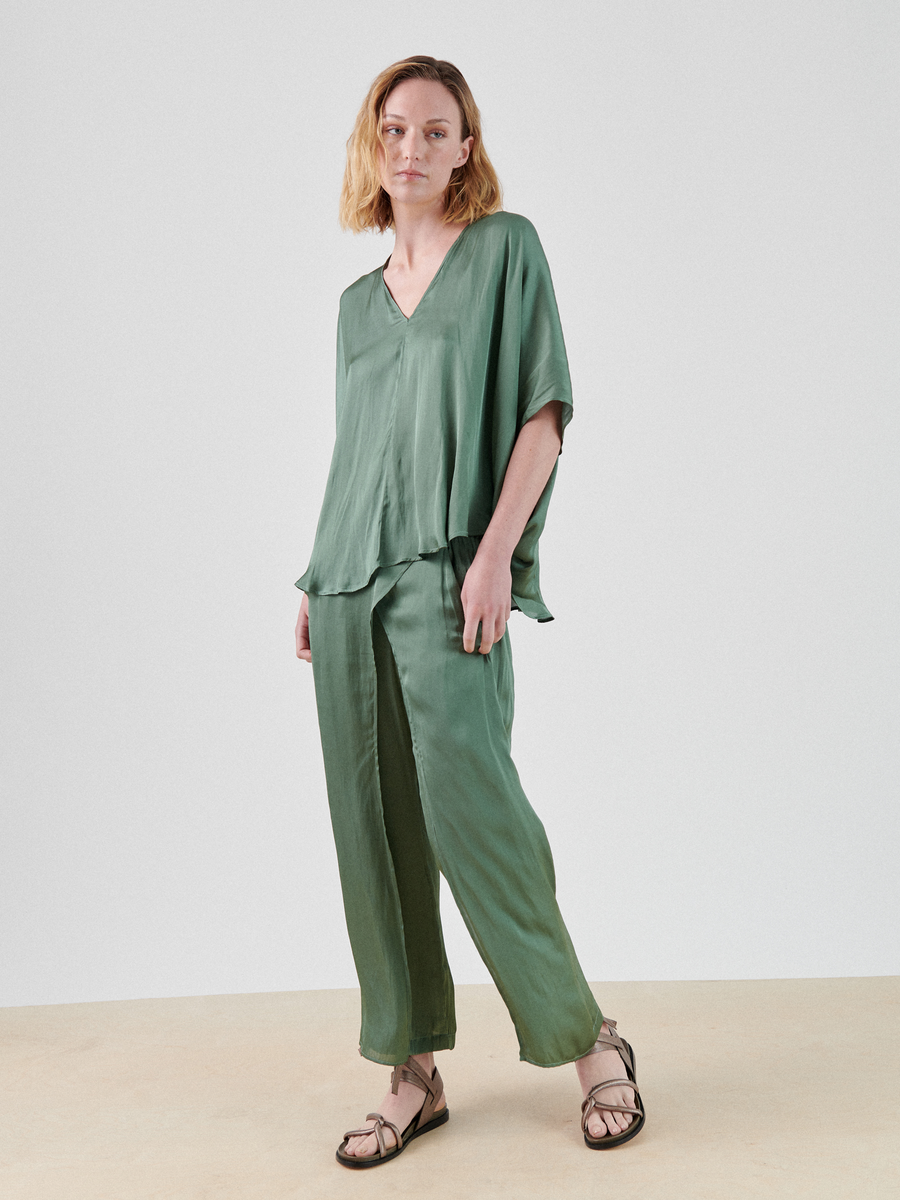 A person with shoulder-length blonde hair is standing against a plain white background, wearing a loose-fitting, green washed georgette Circle Top by Zero + Maria Cornejo with a V-neck and matching green silk wide-legged pants. They are also wearing brown sandals.