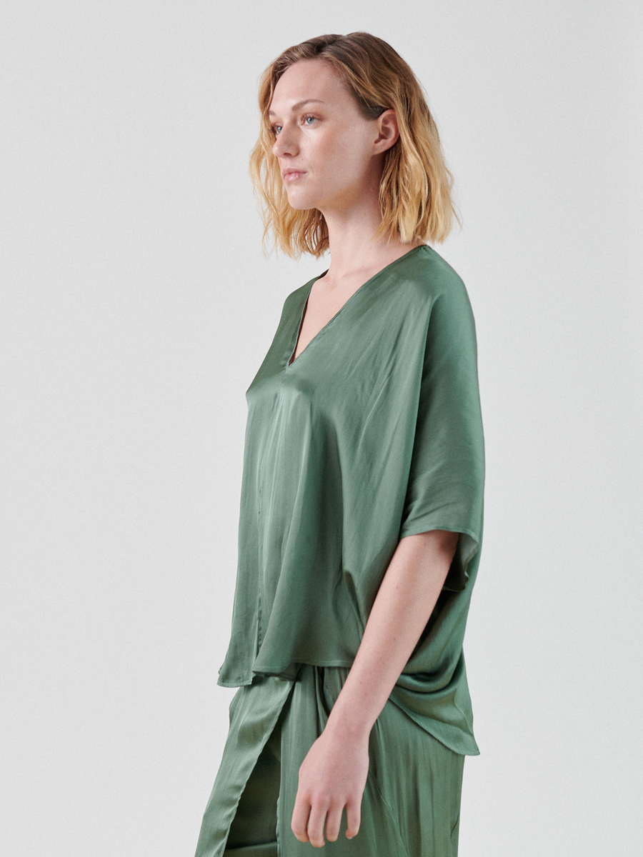 A person with shoulder-length wavy blonde hair is standing in a green, silky outfit made from recycled polyester. The top features a V-neckline and short, loose-fitting sleeves in a washed georgette fabric, while the matching bottom appears to be flowy trousers. This Circle Top from Zero + Maria Cornejo adds an elegant touch to the ensemble. The background is plain and light-colored.