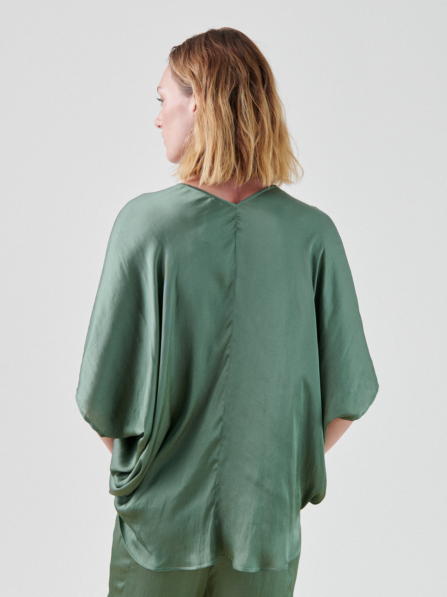 A person with shoulder-length blonde hair stands turned slightly away from the camera, wearing a loose-fitting, green silk Circle Top by Zero + Maria Cornejo with sleeves that reach the elbows. The draped circle silhouette of this washed georgette top features a subtle seam running down the back. The background is plain and light-colored.