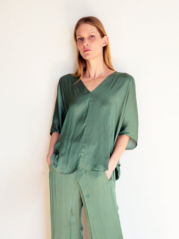 A woman with long blonde hair, standing against a plain white background, wears a loose-fitting, olive-green Circle Top from Zero + Maria Cornejo with elbow-length sleeves and matching wide-leg pants crafted from recycled polyester fabric. Her hands are in her pockets, and she has a neutral expression.