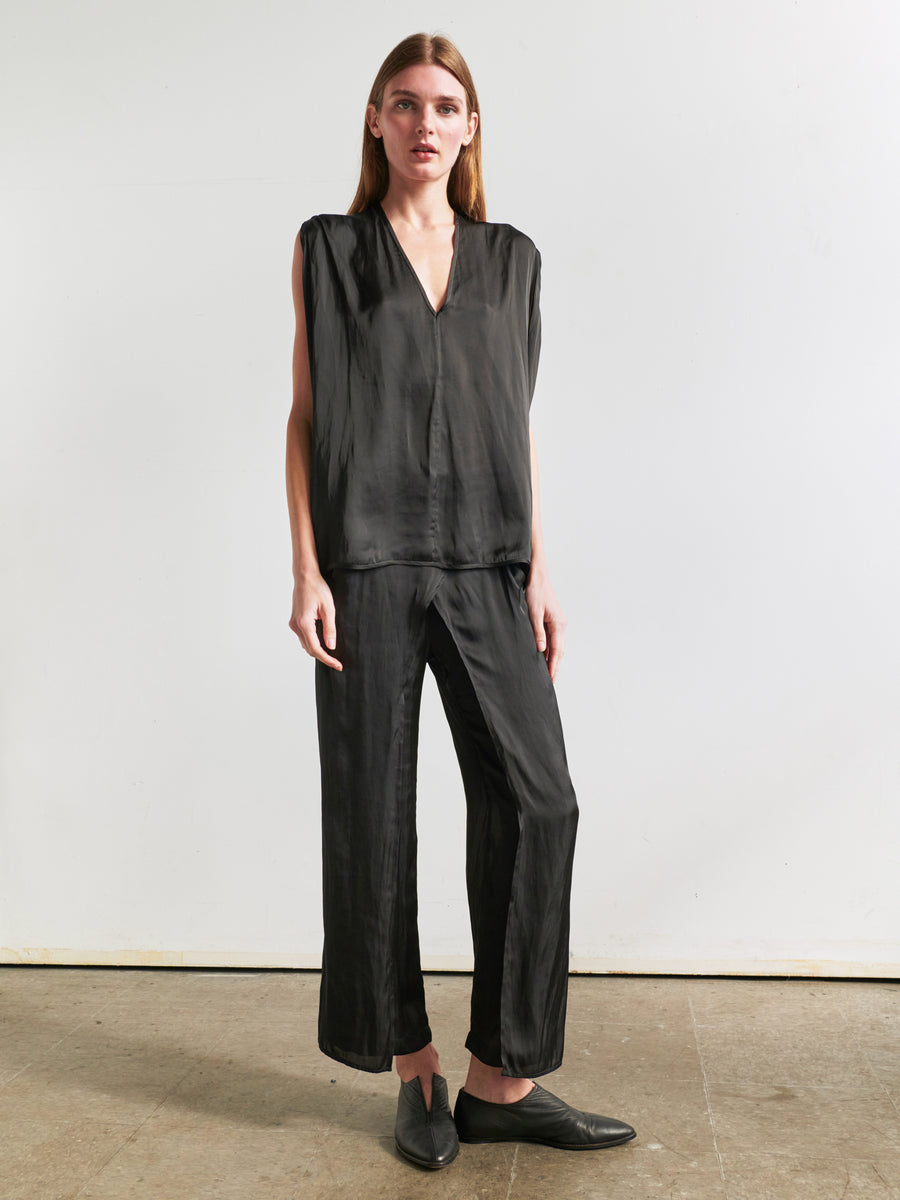 A person with long hair stands against a plain background, wearing the relaxed sleeveless V-neck Ina Tank from Zero + Maria Cornejo, made from recycled polyester and paired with matching black pants. They are also wearing black slip-on shoes. The outfit exudes a sleek, minimalistic style with a draped silhouette.