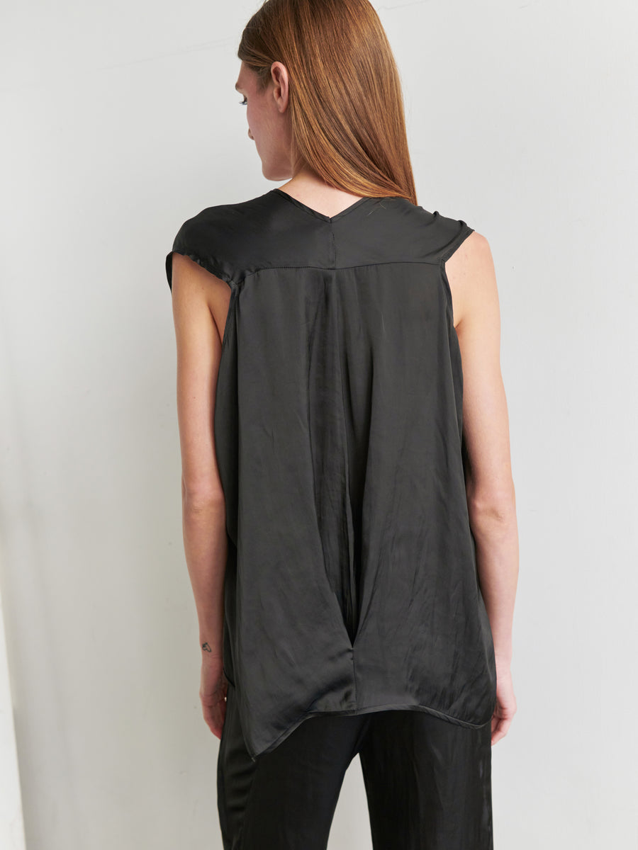 A person with long light brown hair is standing with their back to the camera, wearing the Ina Tank by Zero + Maria Cornejo. This sleeveless v-neck top in recycled polyester features a loose, draped silhouette and is paired with black pants. The background is a plain light-colored wall.