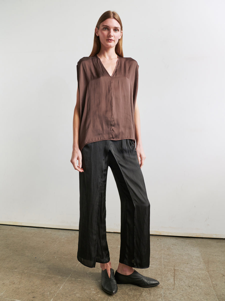 A person with long hair stands against a plain background. They are wearing the Ina Tank by Zero + Maria Cornejo, a loose, brown sleeveless V-neck top, paired with wide-legged black pants to create a draped silhouette. They have a neutral expression and face the camera with their hands relaxed at their sides. Black loafers complete the outfit.