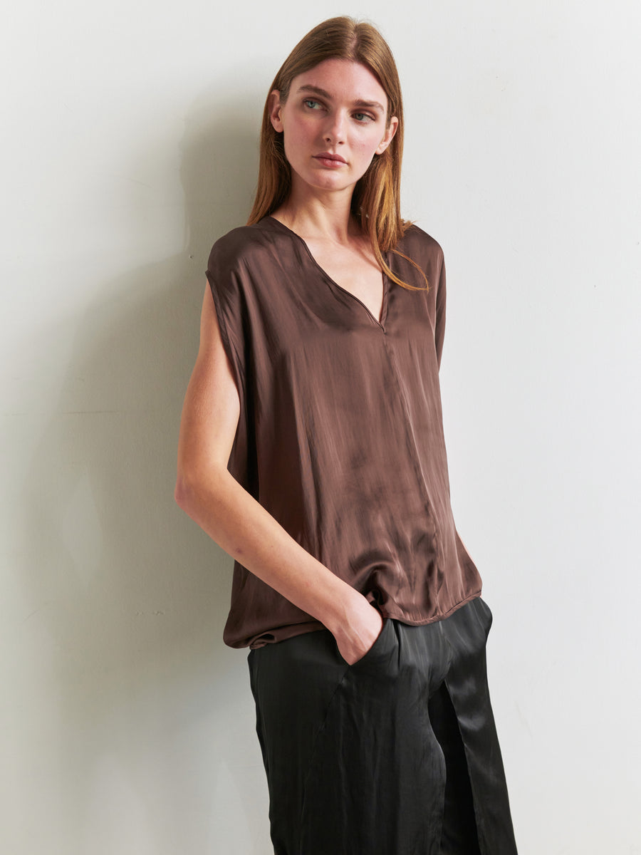 A woman with long brown hair stands against a white wall, wearing the Ina Tank by Zero + Maria Cornejo, made from recycled polyester. She pairs it with dark pants, her hands tucked into her pockets as she gazes thoughtfully to the side, highlighting the draped silhouette of her blouse. The background is neutral and well-lit.