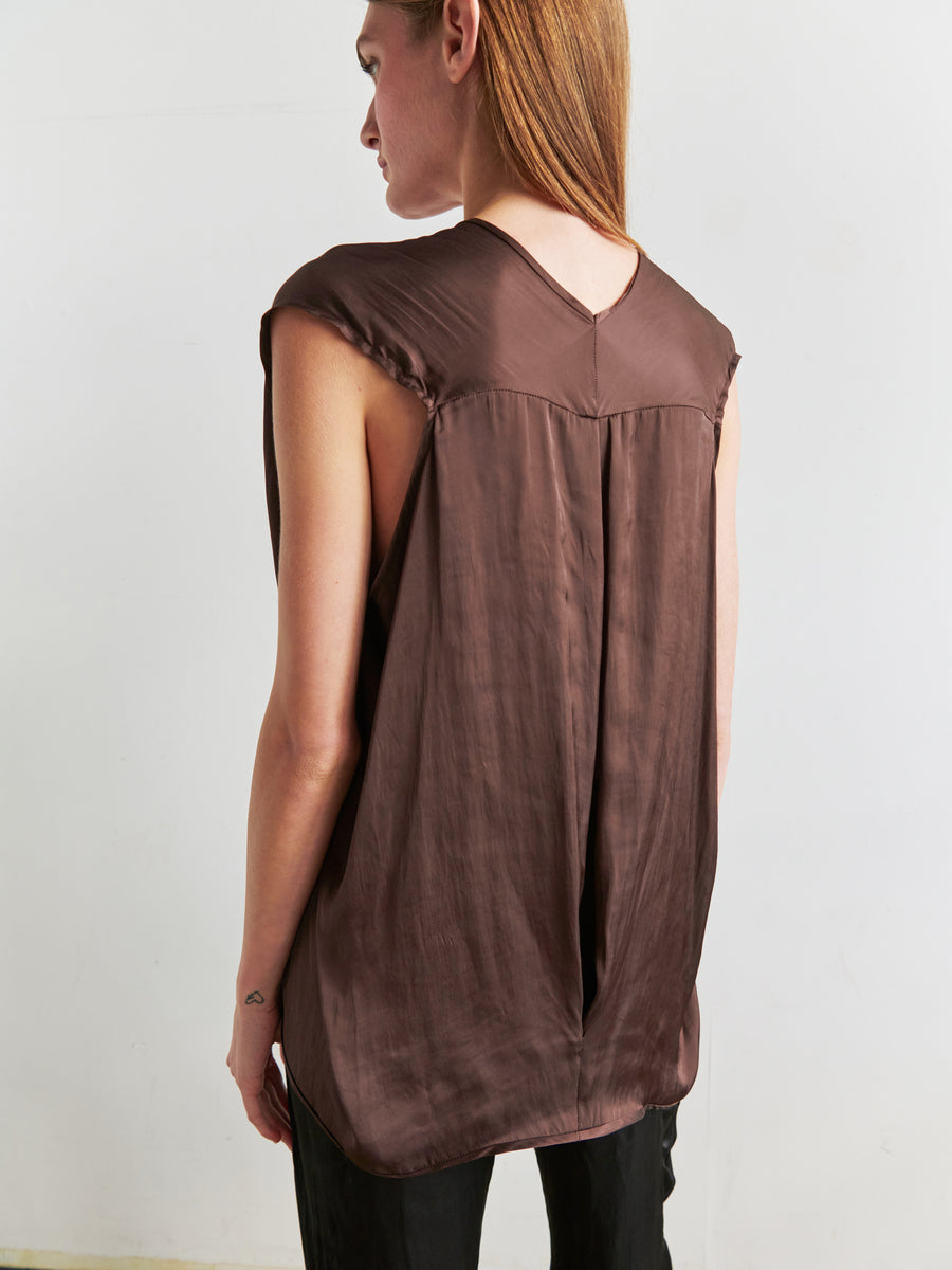 A person with long hair is standing with their back to the camera, wearing the Ina Tank, a sleeveless v-neck top made of silky brown fabric from Zero + Maria Cornejo, featuring a draped silhouette. They pair it with black pants against a plain white wall, offering an elegant yet understated look.