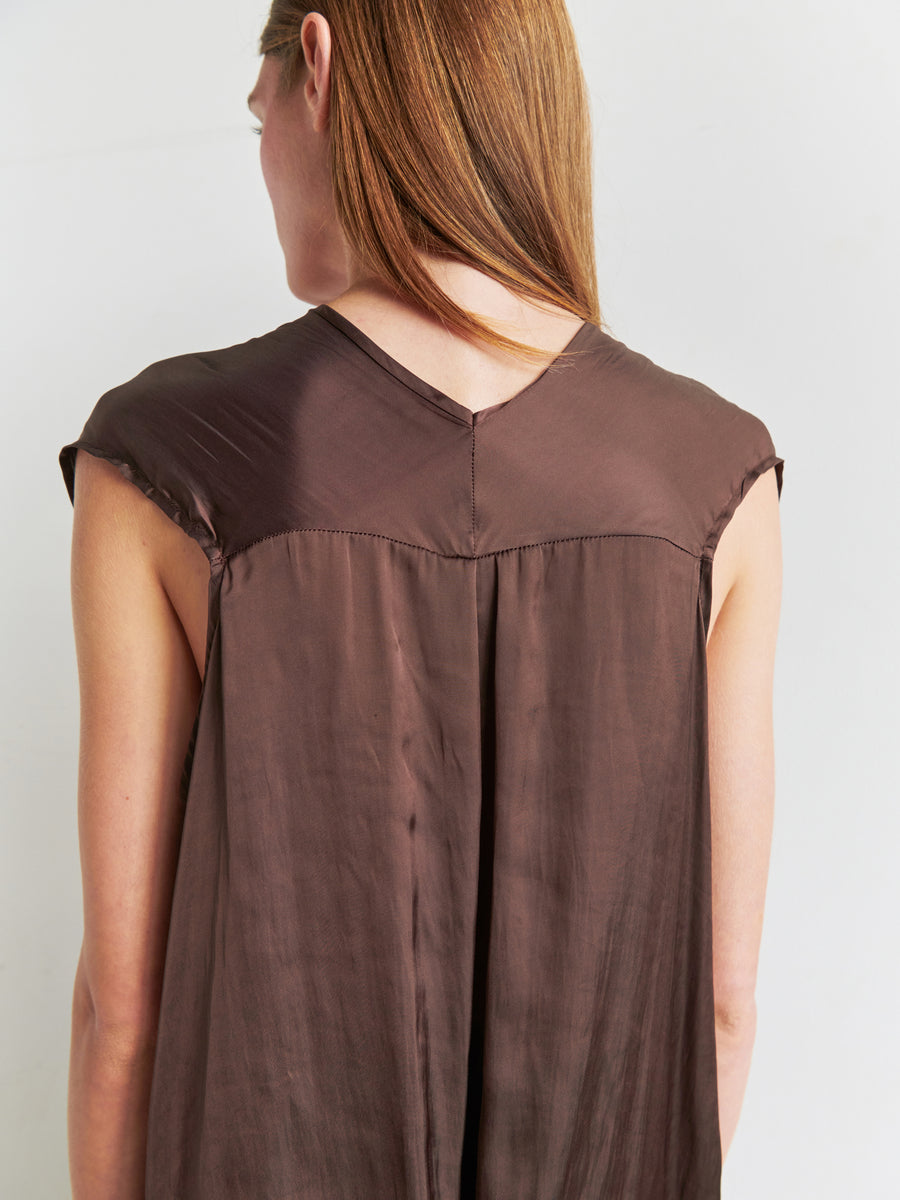 A woman with long, straight hair is seen from the back, wearing the Ina Tank by Zero + Maria Cornejo. The top is a sleeveless v-neck design in a loose-fitting brown style. Crafted from recycled polyester fabric, it appears soft and slightly shiny with a distinctive seam at the back that creates a pleated effect. The background is light and plain.