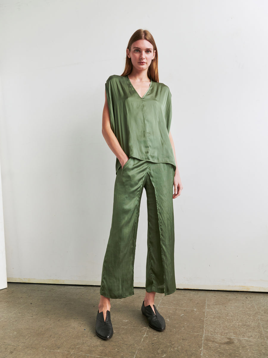 An individual with long hair is standing against a plain wall, wearing an olive green Ina Tank by Zero + Maria Cornejo and matching loose-fitting silk trousers with a draped silhouette. They have one hand in their pocket and are wearing black loafers. The floor is concrete, and the setting appears minimalistic and modern.