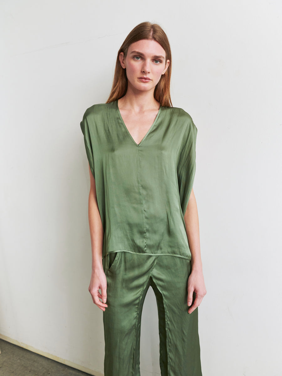 A person with long, brown hair stands against a white wall. They are wearing the Ina Tank by Zero + Maria Cornejo, a loose-fitting green silk outfit featuring a sleeveless V-neck top and matching pants that create a draped silhouette. The person's arms are relaxed at their sides, and they look directly at the camera.