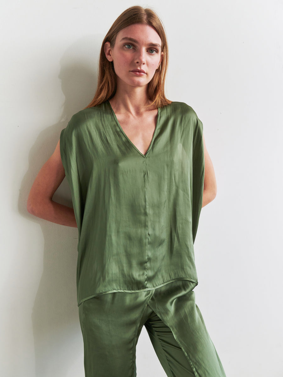 A woman with long, straight hair stands against a white wall, wearing the Ina Tank by Zero + Maria Cornejo. The matching green satin sleeveless v-neck top and loose-fitting pants are made from recycled polyester, creating a draped silhouette. Her arms rest relaxed behind her back as she maintains a neutral expression on her face.