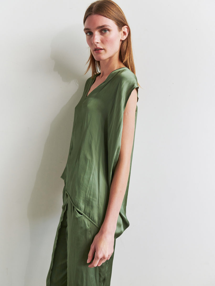 A person with long, straight hair stands against a white wall, wearing a loose, silky green Ina Tank and matching pants made of recycled polyester by Zero + Maria Cornejo. They are turned slightly to the side, looking directly toward the camera with a neutral expression. The draped silhouette adds an elegant touch to their outfit.