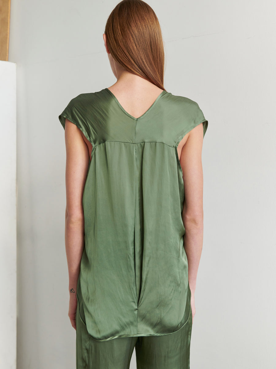 A person with long, straight hair is facing away from the camera, wearing a green Ina Tank in recycled polyester from Zero + Maria Cornejo, paired with matching green pants. The draped silhouette stands out against a minimal and softly lit background.