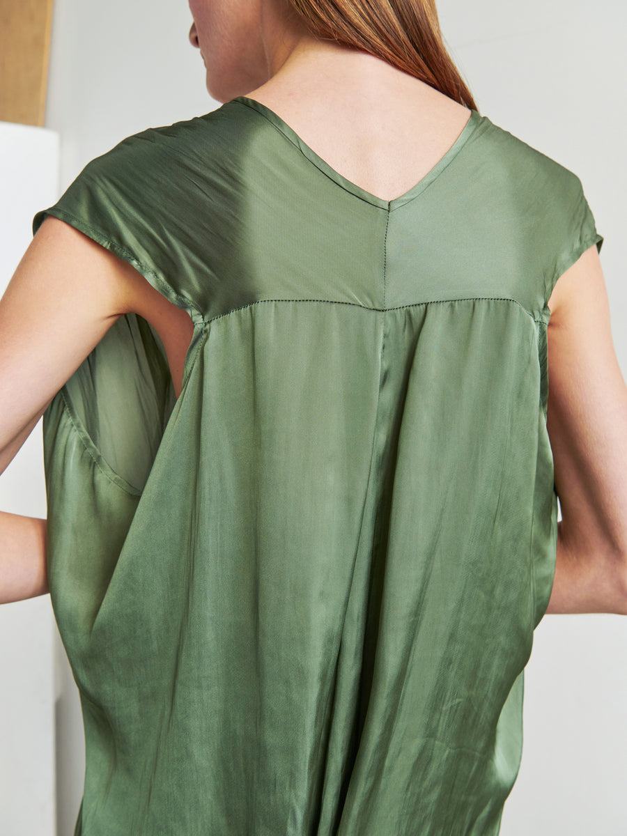 A person with long hair is shown from the back, wearing the Ina Tank by Zero + Maria Cornejo—a shiny, silky green sleeveless v-neck top. The texture and sheen of the recycled polyester fabric are evident, giving a luxurious appearance to the clothing. The draped silhouette adds elegance to the overall look.