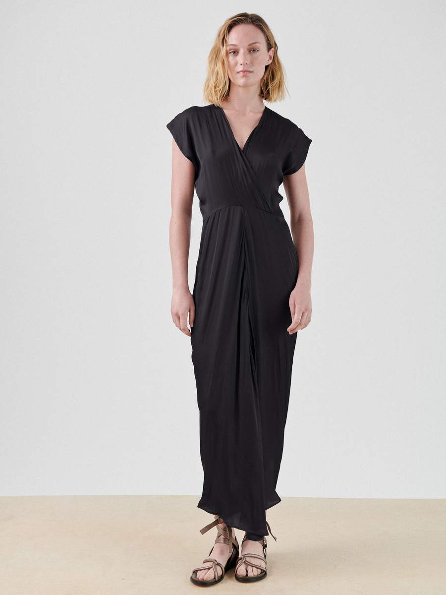 A person with shoulder-length blonde hair is wearing a black, short-sleeved, V-neck Wrap Omi Dress by Zero + Maria Cornejo made from recycled polyester fabric. They are standing against a plain white background and are wearing brown strappy sandals. The dress has a relaxed and flowing appearance.