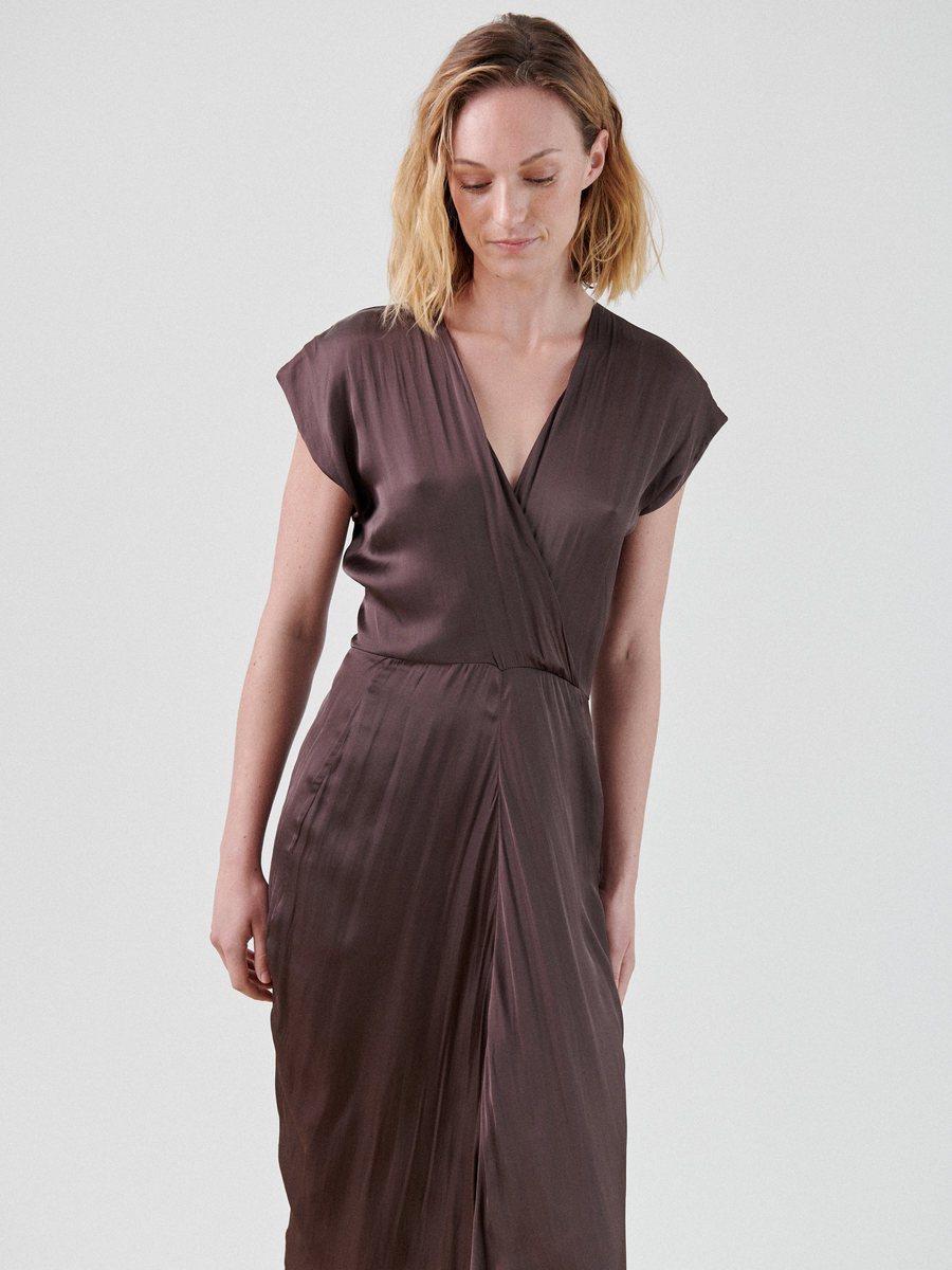 A person with shoulder-length, wavy blonde hair is wearing a silky, brown Zero + Maria Cornejo Wrap Omi Dress with short cap sleeves. They are standing against a plain white background, looking slightly down and to the left with a soft smile.