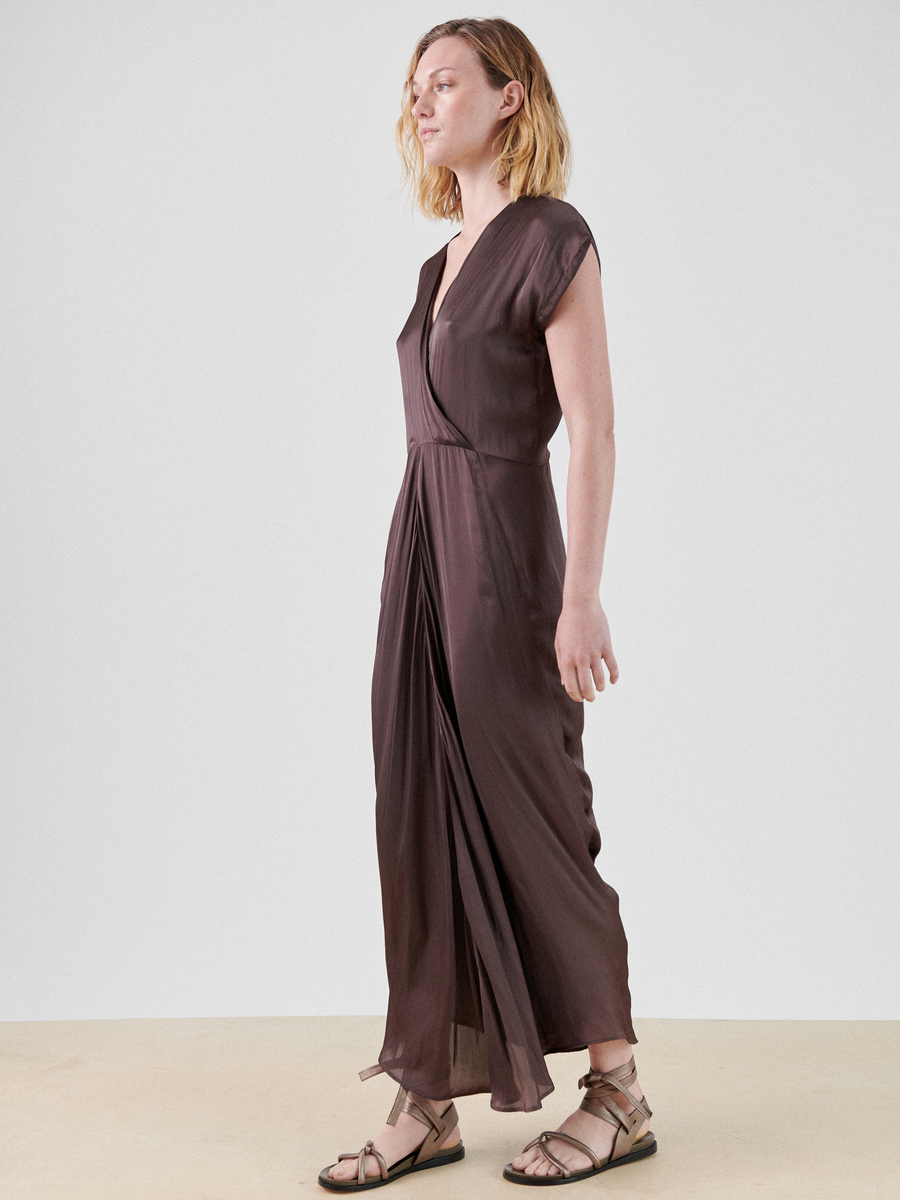 A person with shoulder-length blonde hair is standing against a plain white background. They are wearing a floor-length, sleeveless, deep brown Wrap Omi Dress by Zero + Maria Cornejo made of recycled polyester fabric and beige sandals. The person appears to be looking slightly upward and to the right.