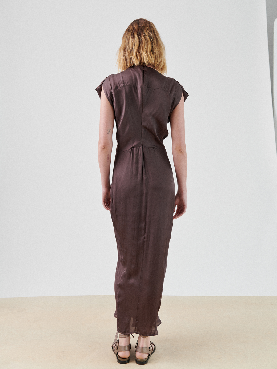 A woman with blonde hair, seen from the back, is wearing a short-sleeved, ankle-length brown Wrap Omi Dress by Zero + Maria Cornejo. She stands indoors on a light-colored floor, facing a plain white wall. She has a small tattoo on her right arm and is wearing brown sandals.