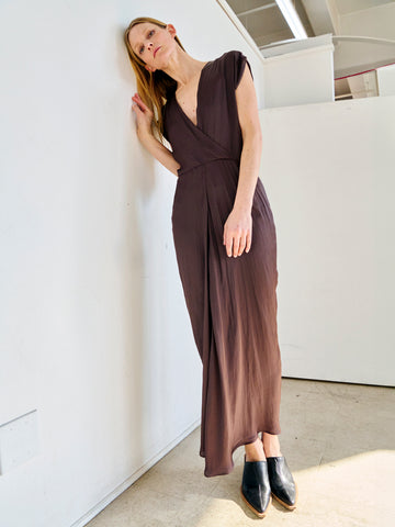 A woman with blonde hair is leaning against a white wall. She is wearing a Wrap Omi Dress by Zero + Maria Cornejo with short sleeves and black shoes. Tilting her head slightly to the side, her right hand touches her hair. The setting appears to be indoors with natural lighting.