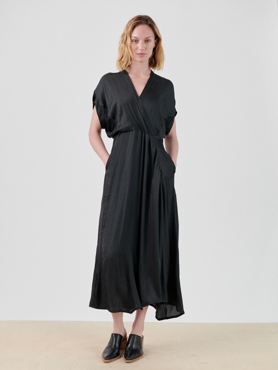 A person with shoulder-length blonde hair is standing against a plain background, wearing a black, short-sleeved, wrap-effect V-neck Long Aki Wave Dress made of recycled polyester with a cinched waist by Zero + Maria Cornejo. They have their hands in their dress pockets and are wearing black slip-on shoes.