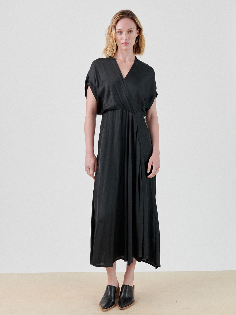 A woman with blonde hair stands against a plain white background, wearing a black, ankle-length Long Aki Wave Dress by Zero + Maria Cornejo featuring short sleeves and a wrap-effect v-neckline with pleats. She is also wearing black slip-on shoes.