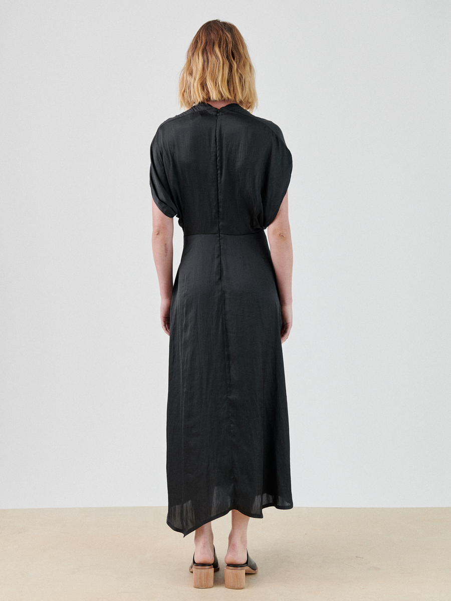 A person with shoulder-length hair, wearing a lightweight georgette dress and beige shoes, is standing and facing away from the camera. The ankle-length Long Aki Wave Dress by Zero + Maria Cornejo features a wrap-effect v-neck and short sleeves. The background is plain and light-colored.