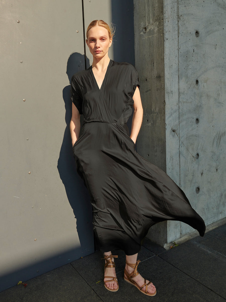 A person with light hair is standing against a wall, wearing a lightweight georgette Long Aki Wave Dress by Zero + Maria Cornejo in black and brown sandals. The lighting creates a sharp shadow on the wall, and the dress appears to be made of soft, washable fabric. The person has their hands in their pockets.