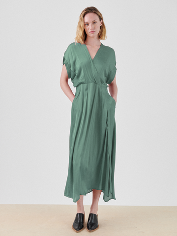 A person with shoulder-length blonde hair stands against a plain white background, wearing the Long Aki Wave Dress by Zero + Maria Cornejo, a flowing green lightweight georgette dress with an asymmetrical skirt and short sleeves. Black slip-on shoes complete the look. Their hands are in the dress pockets, and they have a neutral expression.